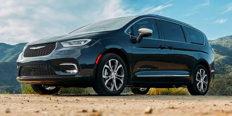 A dark blue Chrysler Pacifica Pinnacle minivan parked on a dirt road with mountains and sky in the background.