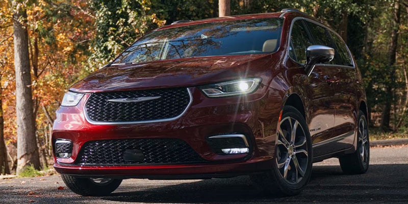 A shiny cherry-red Chrysler Pacifica minivan with bright headlights on a road, with autumn trees in the background.