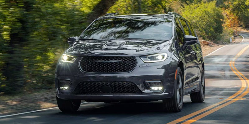 Front view of a dark gray Chrysler minivan driving on a winding road with trees in the background.