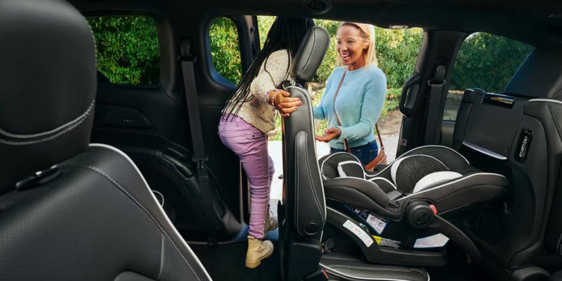 A woman helps a child enter the back seat of a car equipped with a baby car seat.