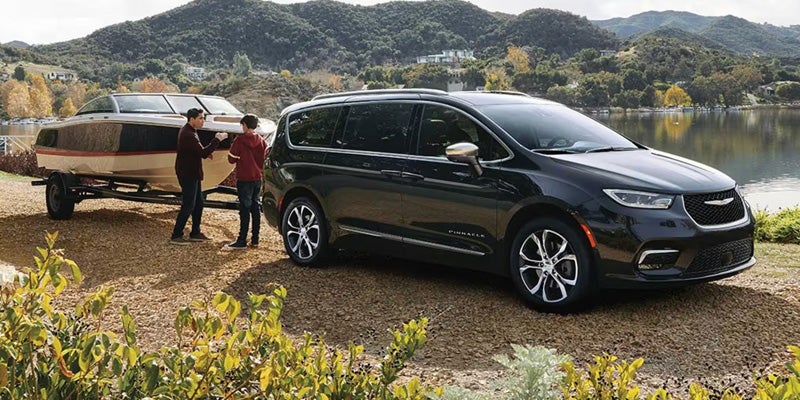 Black minivan on a gravel shore next to a lake, towing a boat, with two people preparing it.