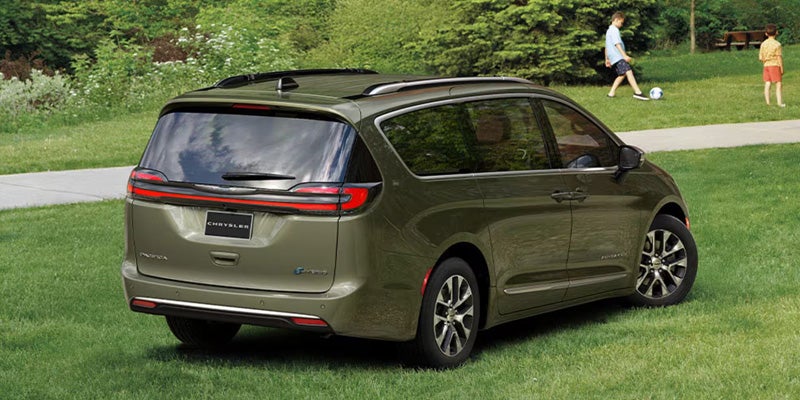 Rear view of a dark olive green Chrysler Pacifica minivan with chrome accents, parked on green grass. Two children play in the background.