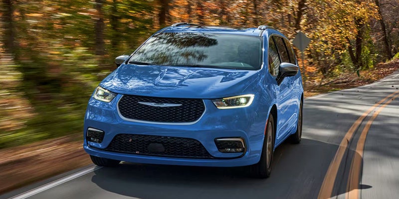 Blue minivan drives fast on a winding road bordered by autumn trees; motion blur suggests speed.
