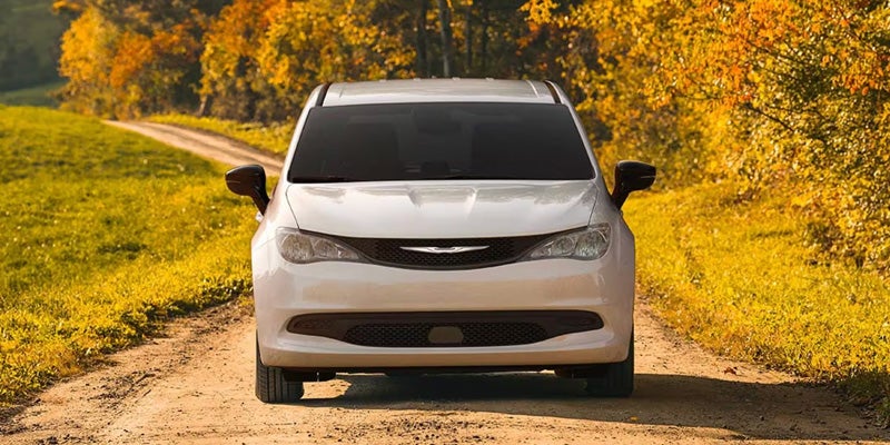 A white Chrysler Pacifica minivan on a dirt road, surrounded by green and golden autumn foliage.