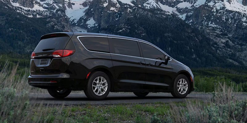 A black minivan parked on an asphalt road with a snow-capped mountain range and evergreen trees in the background.