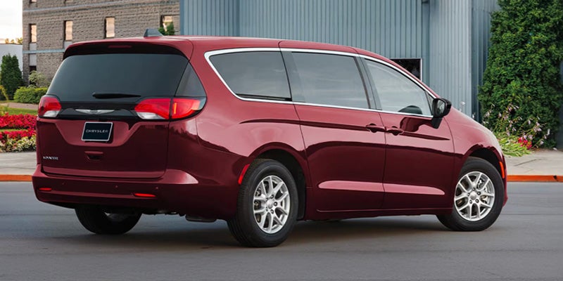 Rear driver-side view of a dark red Chrysler Pacifica minivan parked on asphalt next to a building.