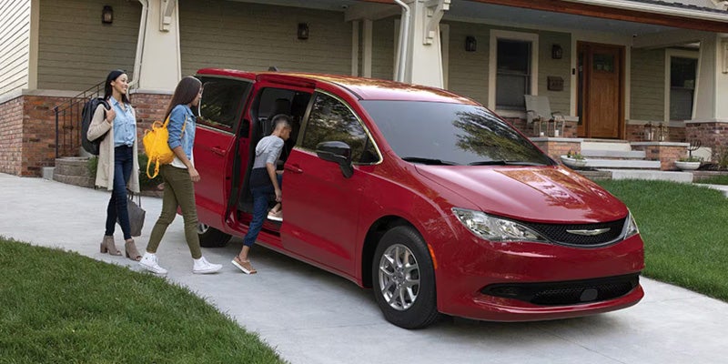 A family of three (two women, one boy) getting into a red minivan parked in a driveway in front of a house.