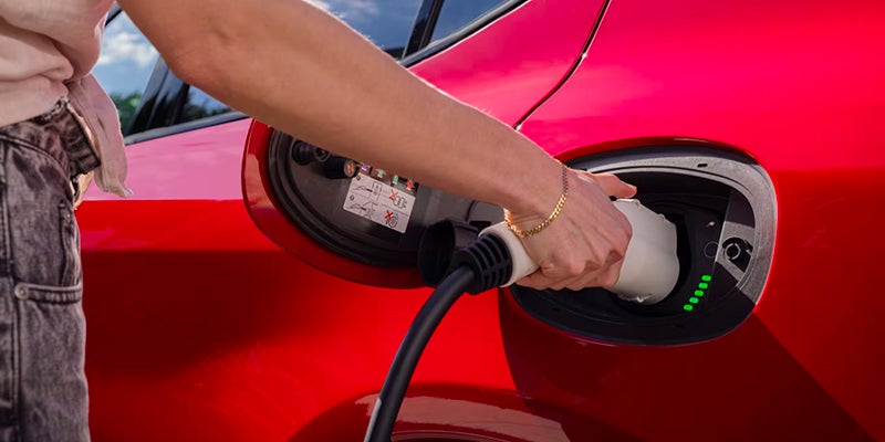 A person's hand plugging a white charging cable into the charging port of a red electric car, with green indicator lights showing.