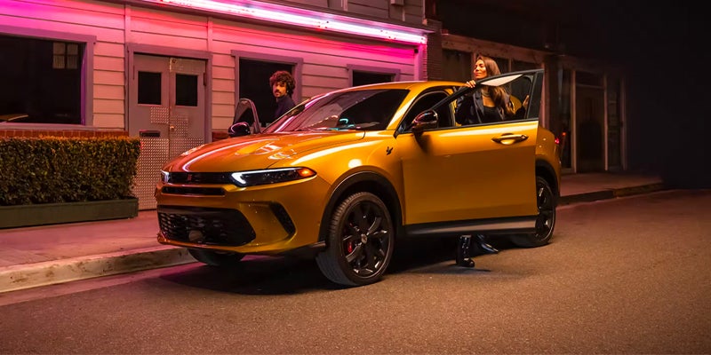 A man and a woman getting into or out of a shiny gold new Dodge Hornet car at night outside a building.