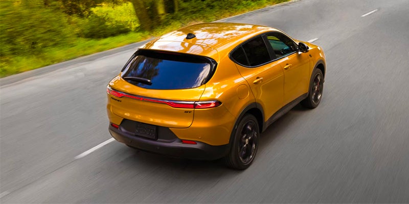 Rear overhead view of a gold Dodge Hornet GT SUV driving on a winding gray road with blurred green trees.