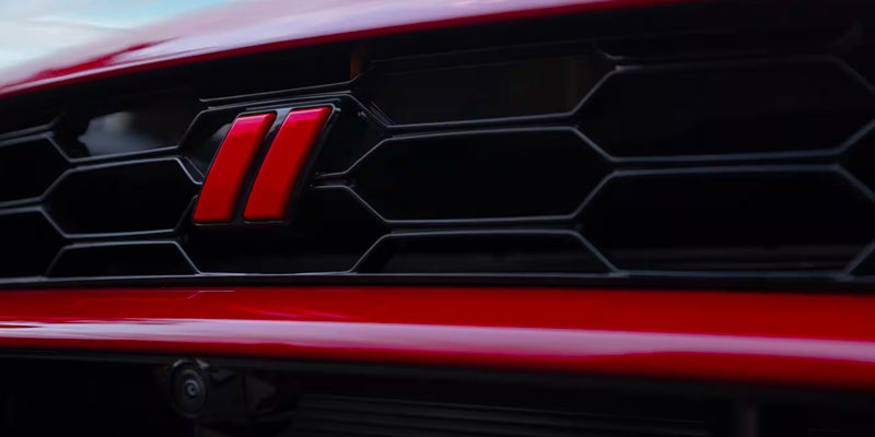 Close-up of a red car's lower grille with a prominent, bright red "II" emblem on a dark hexagonal mesh.