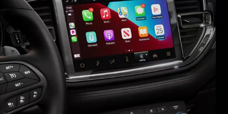 A car's interior showing a steering wheel on the left and a large infotainment screen displaying various app icons.