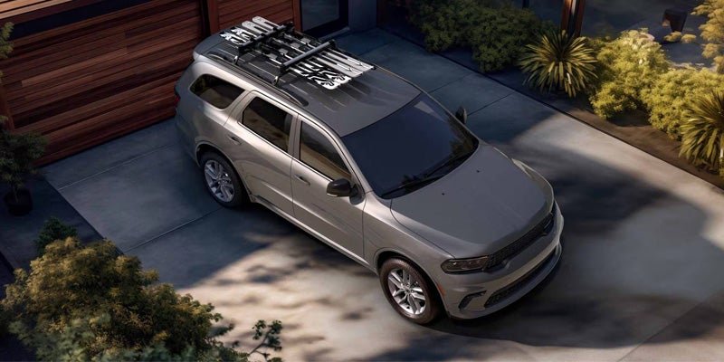 Gray SUV with skis on roof rack parked on a driveway next to a wooden garage door and green landscaping.