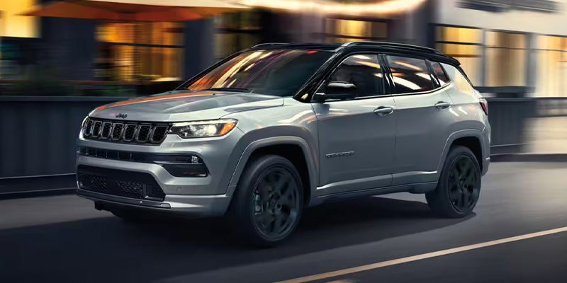 Gray Jeep Compass SUV with a black roof drives quickly down a city street at dusk.