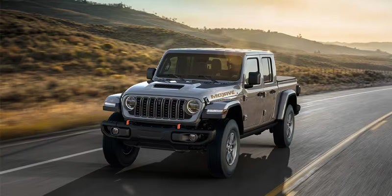 A gray Jeep Gladiator Mojave pickup truck drives on a paved road through a dry, hilly landscape at sunset