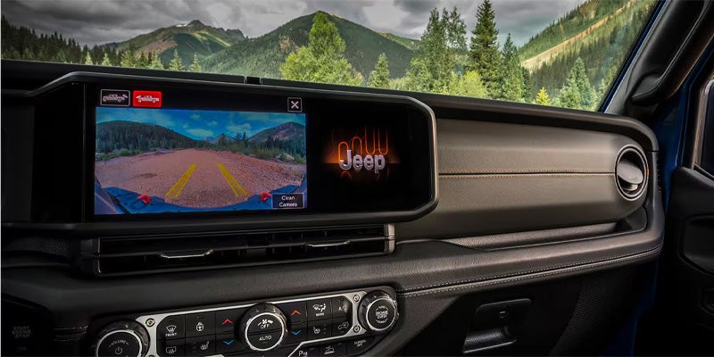 A Jeep Wrangler's infotainment screen displays a reverse camera view of a dirt trail in front of mountains and trees.