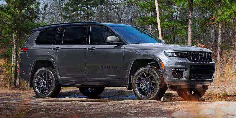 Dark gray Jeep Grand Cherokee parked on a rocky path in a forest with tall green trees.