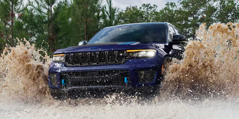 Dark blue Jeep Grand Cherokee 4xe with blue recovery hooks drives through muddy water, splashing high on both sides.