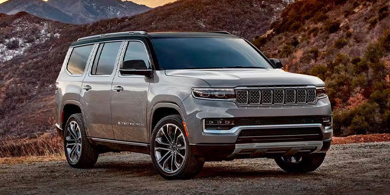 A gray Jeep Grand Wagoneer SUV with a black roof parked on a dirt road in front of mountains.