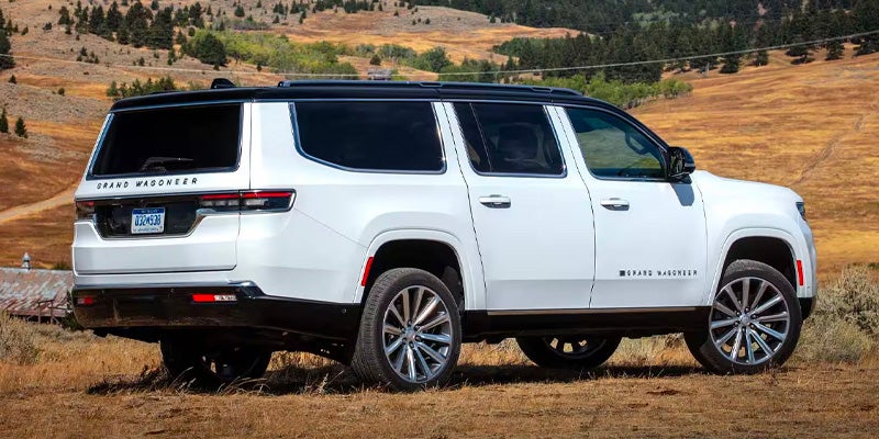 Rear quarter view of a white Grand Wagoneer SUV parked on a dry, grassy hillside with trees in the background.