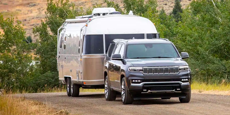 A dark grey SUV towing a silver Airstream trailer on a dirt road, surrounded by green trees and dry grass.