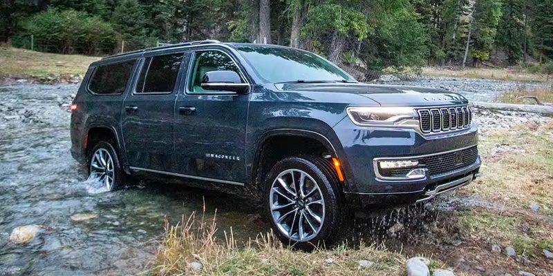 A dark blue Wagoneer SUV driving through a shallow stream in a forest, splashing water as it goes.