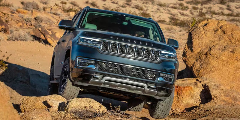 A dark grey SUV, a Jeep Wagoneer, navigating a rocky, dirt terrain uphill from a front-facing perspective.