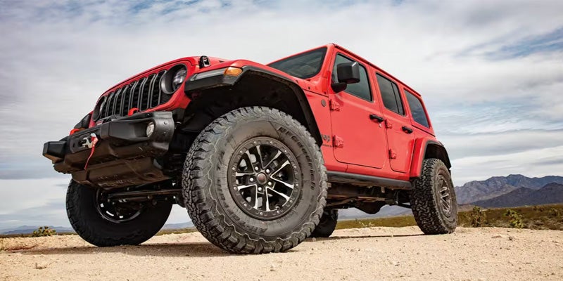 A bright red 4-door Jeep Wrangler with large off-road tires, parked on a dirt road with mountains in the background.