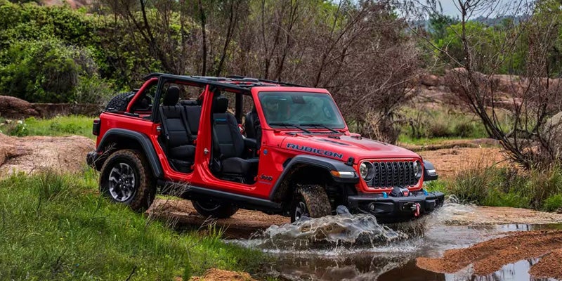 A red Jeep Rubicon, with doors removed, drives through a shallow stream in a rocky, wooded landscape.