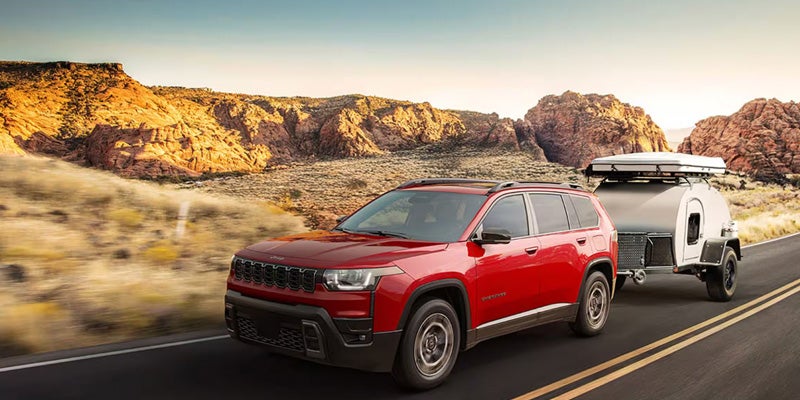Red Jeep Commander SUV towing a teardrop camper on a desert highway with rocky hills in the background.