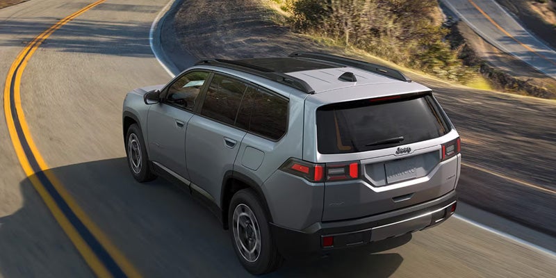 Rear view of a light gray Jeep minivan driving on a curved road, surrounded by dry vegetation.