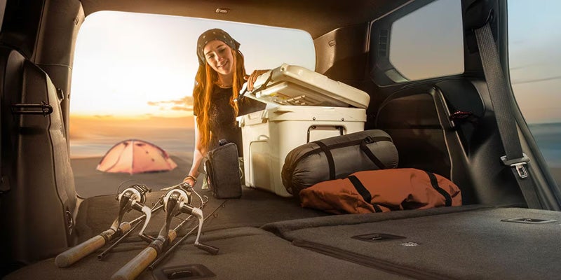 A woman loading camping gear and fishing rods into the back of an SUV at sunset on a beach with a tent.