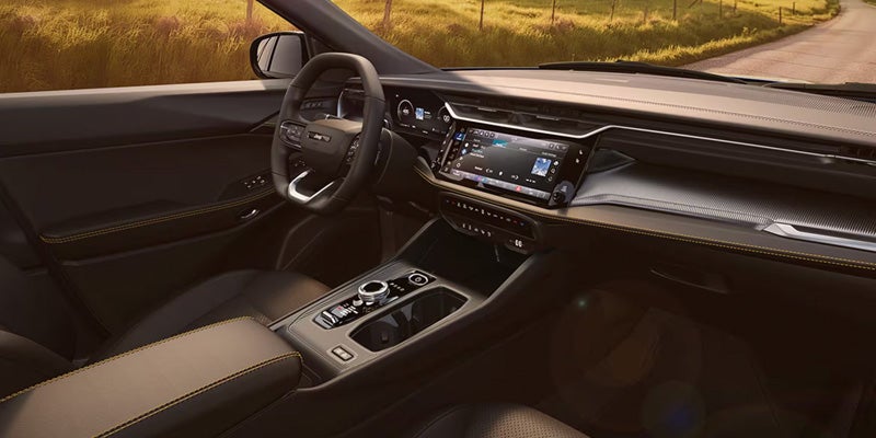 Interior view of a car with a black steering wheel, infotainment screen, and light brown armrest with yellow stitching.