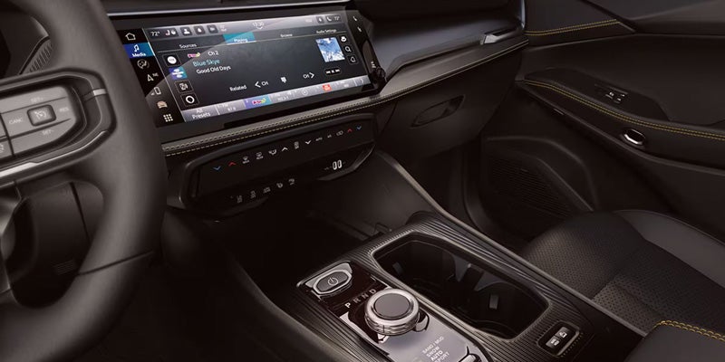 The dashboard and center console of a car, featuring a large infotainment screen playing music, climate controls, and a rotary gear selector.