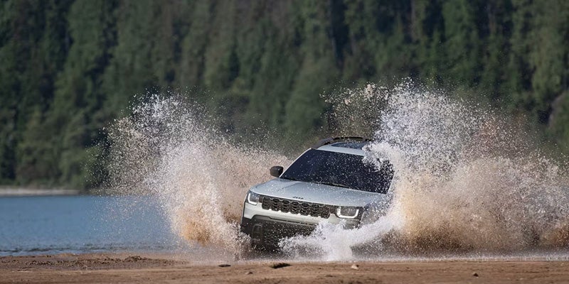 A white Jeep Compass splashing through muddy water on a dirt path with trees and a lake in the background.
