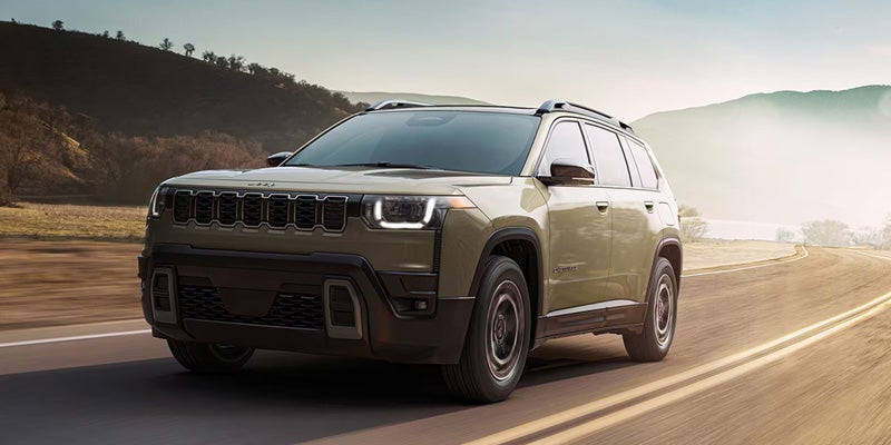 A light green Jeep Commander drives on a winding road with mountains and hazy skies in the background.
