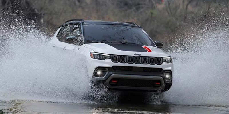 White Jeep Compass with a black roof and red stripe on the hood driving through a large puddle, splashing water.