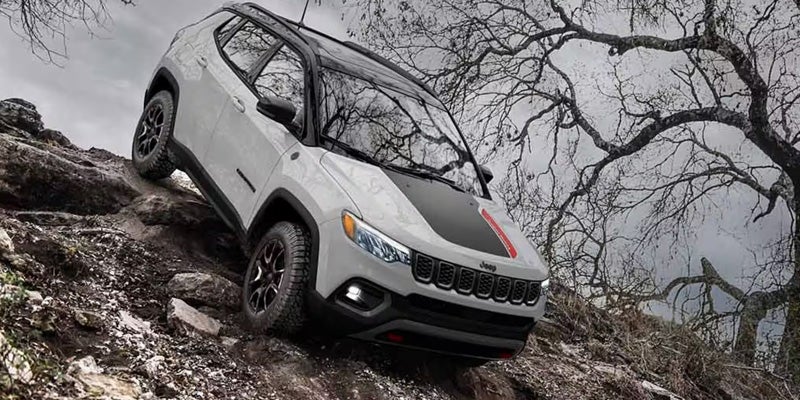 A gray Jeep Compass Trailhawk with a black roof and red hood stripe drives up a rocky, dirt hill.
