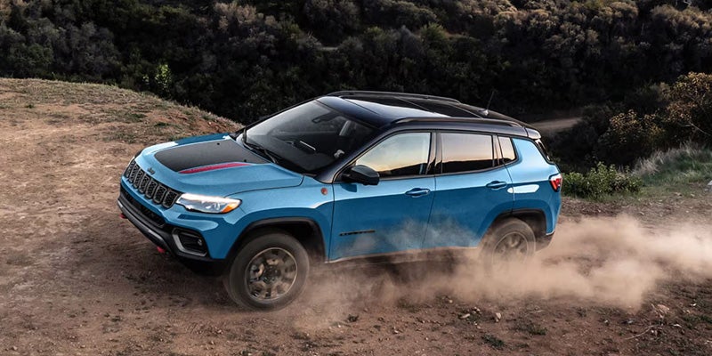A blue Jeep Compass Trailhawk with a black roof kicks up dust while driving on a dirt road.