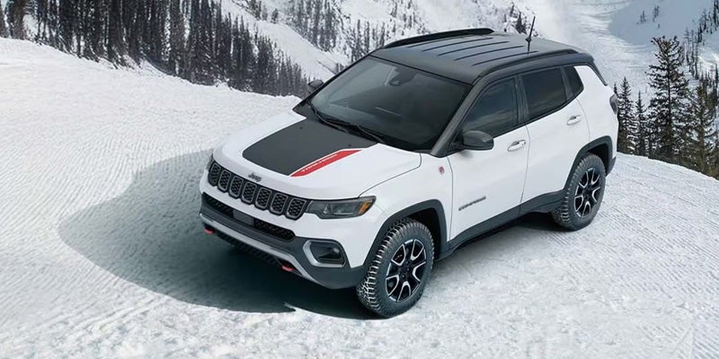 A white Jeep Compass with a black hood and roof is parked on a snowy slope with pine trees in the background.
