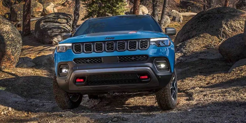 A blue Jeep Compass Trailhawk parked on a rocky off-road trail with sunlight filtering through trees.