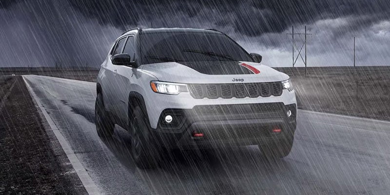 A white Jeep Compass with a black hood stripe and red accents drives on a wet road in heavy rain.