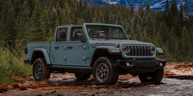 Slate blue Jeep Gladiator Rubicon pickup truck partially submerged in a shallow rocky river within a pine forest.