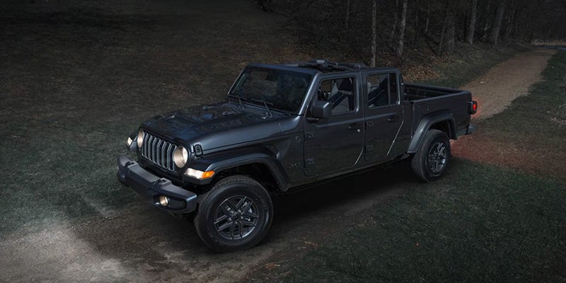 A dark grey Jeep Gladiator pickup truck parked on a grassy path with its headlights illuminating the path ahead
