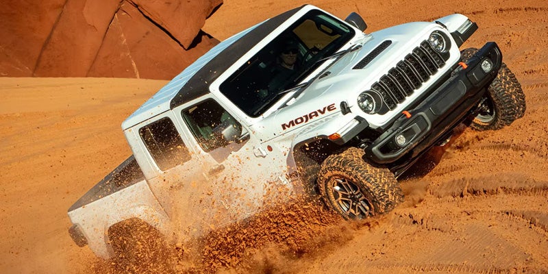A white Jeep Gladiator Mojave pick-up truck drives on an angle up a steep, sandy hill, kicking up dirt.