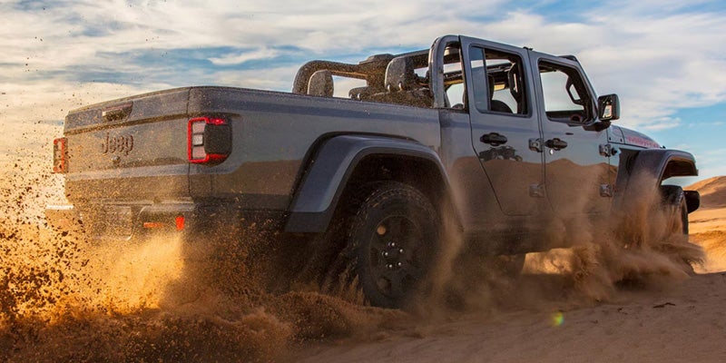 Dark grey Jeep Gladiator truck kicking up sand and dust while driving through a desert landscape under a clear sky.