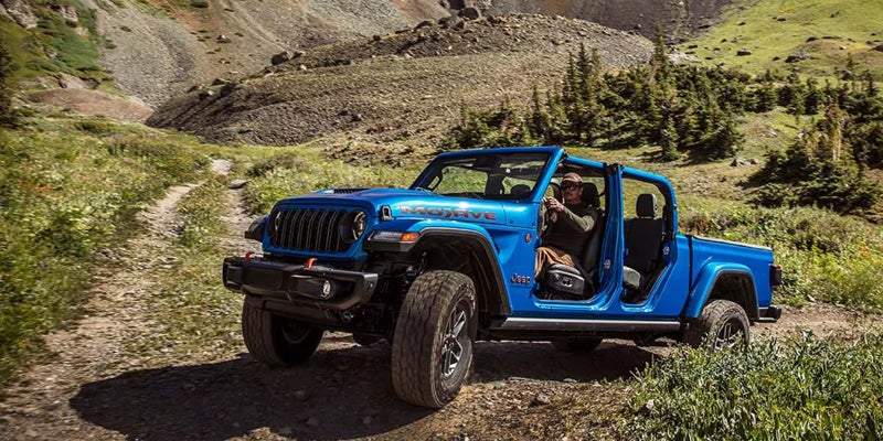 A blue Jeep Gladiator Mojave, with doors removed, driven by a man, on a dirt path in a rocky, treed landscape