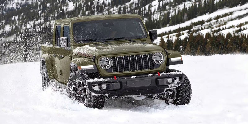 A green Jeep Gladiator Rubicon pickup truck drives through deep snow, kicking up white powder against a snowy mountain backdrop.