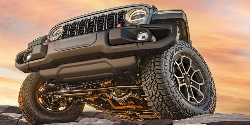 A gray Jeep Wrangler, seen from a low angle, driving over rocks against an orange and blue sunset sky.