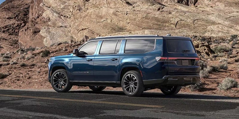 Rear view of a dark blue Grand Wagoneer L SUV driving on a desert road with rocky hills in the background.